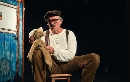 Der ältere Schauspieler sitzt vor dem Holzhaus und hat einen hellen Plüschhasen auf seinem Schoss mit dem er spricht.