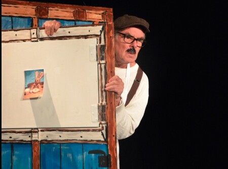 Der ältere Schauspieler schaut vorsichtig hinter einem Holzhaus hervor. Er hat einen Schnauz, eine Brille und eine Mütze an.