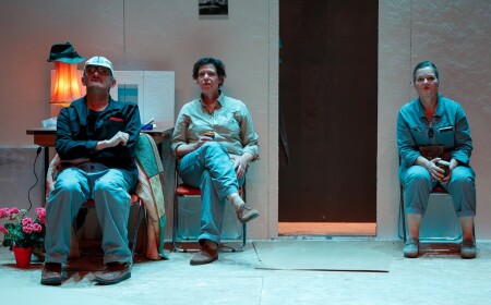 Alle drei Schauspieler sitzen still auf einem Stuhl in einem fast leeren Raum.