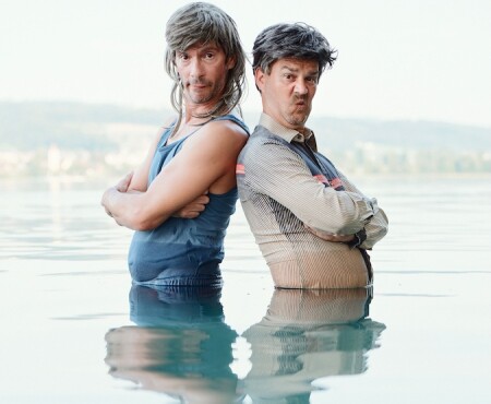 Die beiden Männer stehen Rücken an Rücken immer noch im Wasser und machen erneut ein komisches Gesicht.