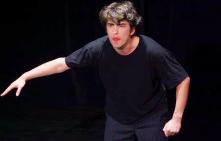 Das Bild zeigt einen jungen Schauspieler. Er steht leicht gebckt, blickt ins Publikum, hat rote Lippen und dunkles Haar.