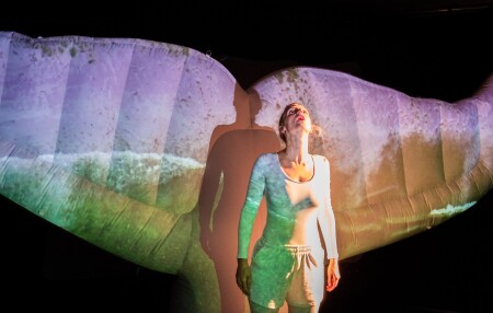 Die Performerin steht auf der Bühne, hinter ihr eine riesige Walflosse. Das Licht ist in grün, lila und gelb, sehr künstlich.